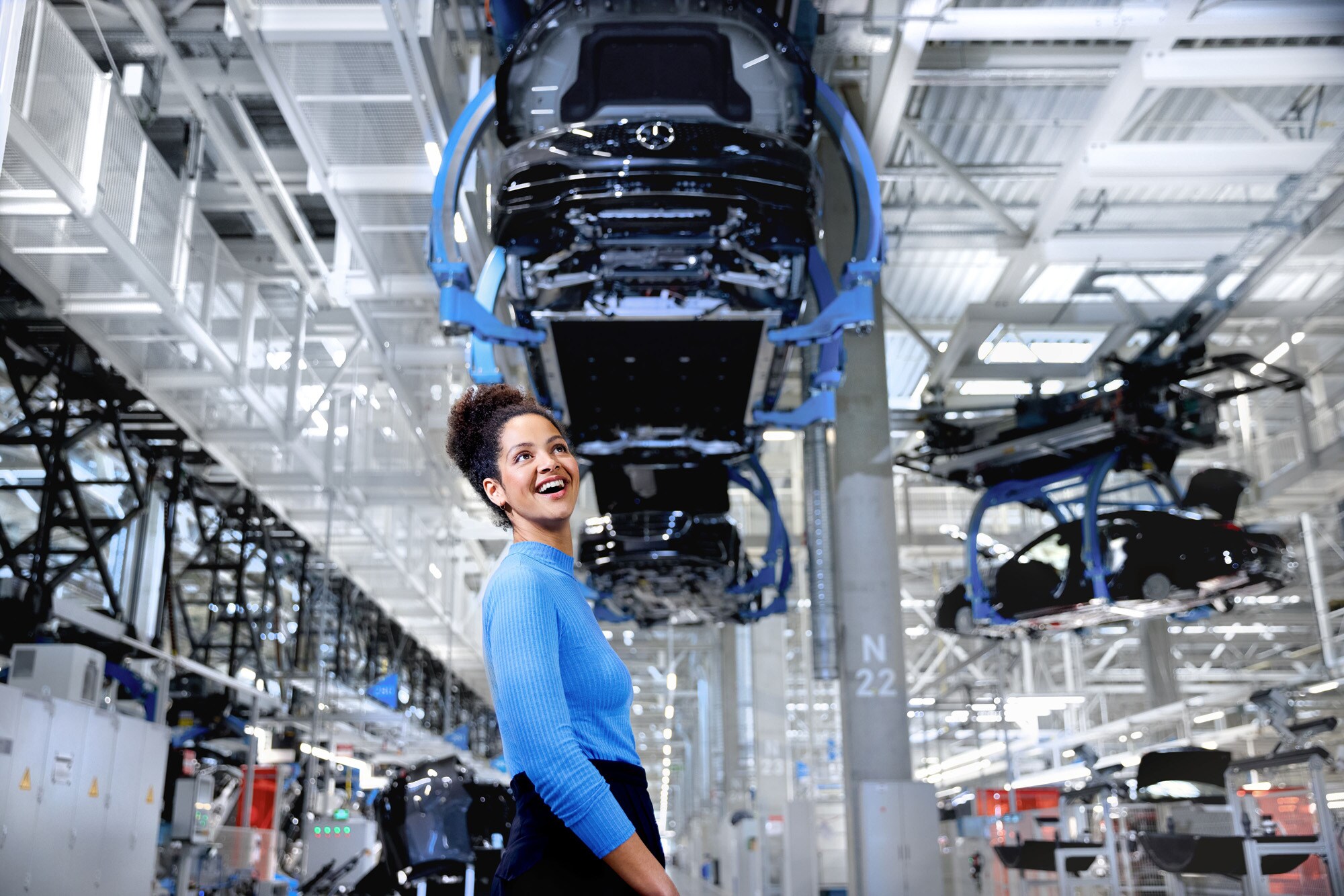 Werksabholung | Mercedes-Benz. Eine Frau steht lachend neben einem Fahrzeug auf einer Hebebühne in einem Mercedes-Benz Werk.