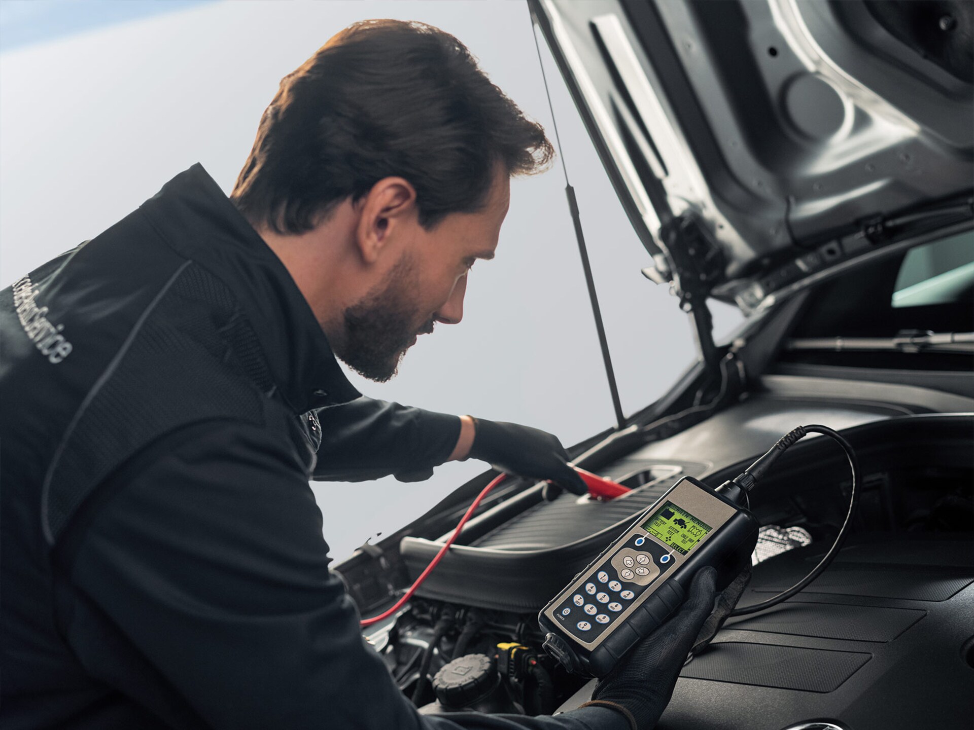 Wintersaktion | Mercedes-Benz Ein junger Mercedes-Benz Mechaniker mit blauem Arbeitsanzug und Handschuhen prüft die Batterie eines Fahrzeugs.