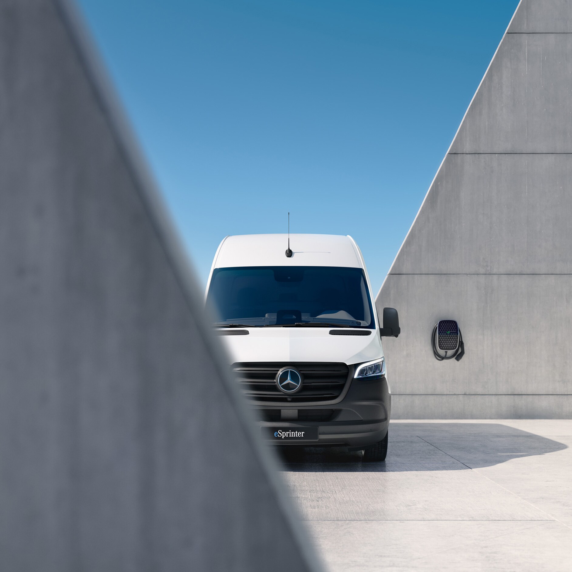 Mercedes-Benz | eSprinter Kastenwagen Ein vollelektrischer eSprinter in Frontansicht vor und hinter einer grauen Mauer. Im Hintergrund eine Wallbox.