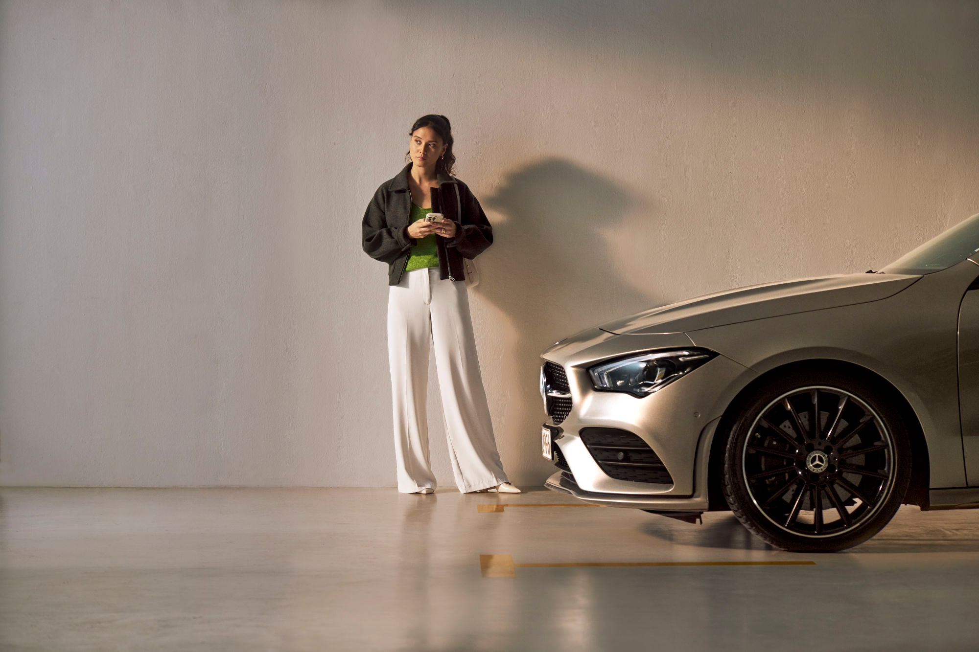 Service VorteilsProgramm | Service Select | Mercedes-Benz. Eine junge Frau steht vor einem Mercedes-Benz Fahrzeug mit Ihren Handy in der Hand und blickt nachdenklich.