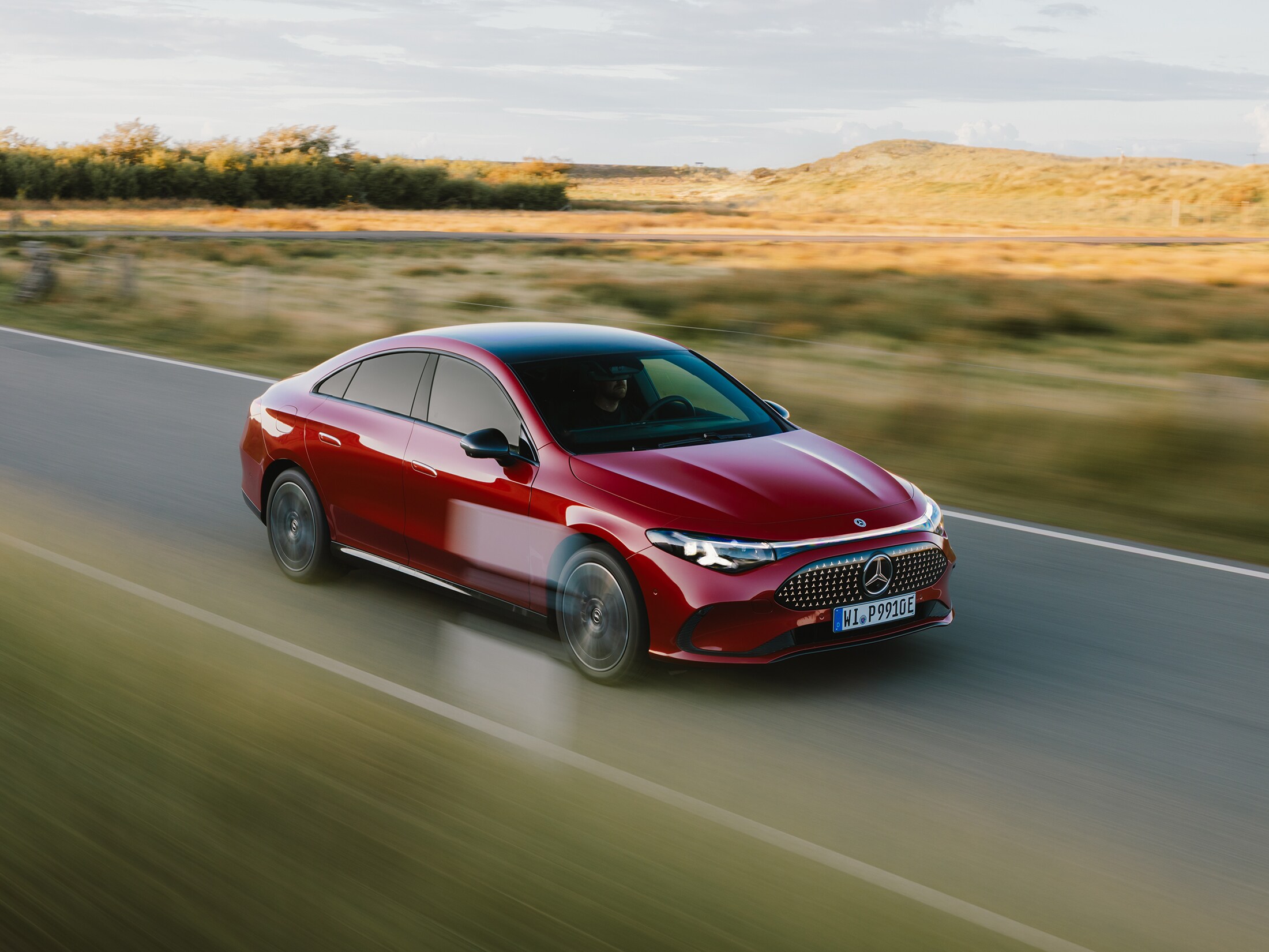 Mercedes-Benz x Sylt | Mercedes-Benz. Ein roter elektrischer CLA fährt auf einer Straße zwischen weiten Grasfeldern auf Sylt.