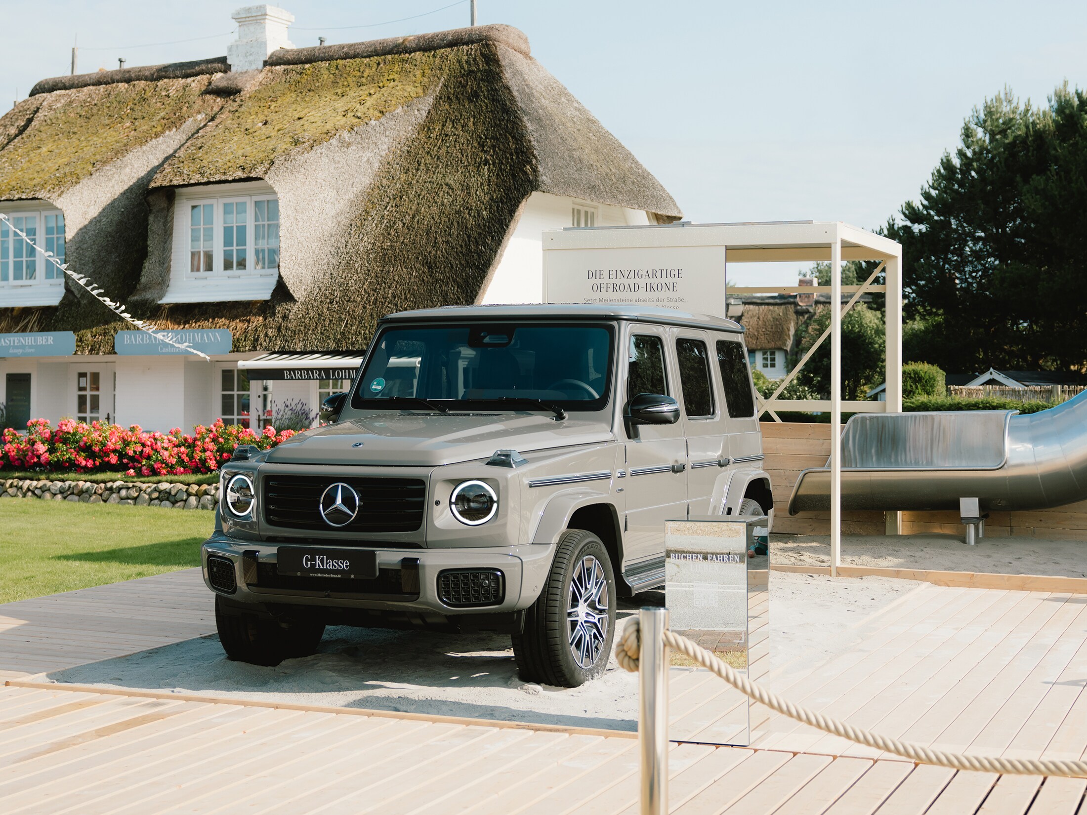 Mercedes-Benz x Sylt | Mercedes-Benz. Eine graue G-Klasse steht auf einer hölzernen Ausstellungsfläche vor einem Haus mit Rieddach.