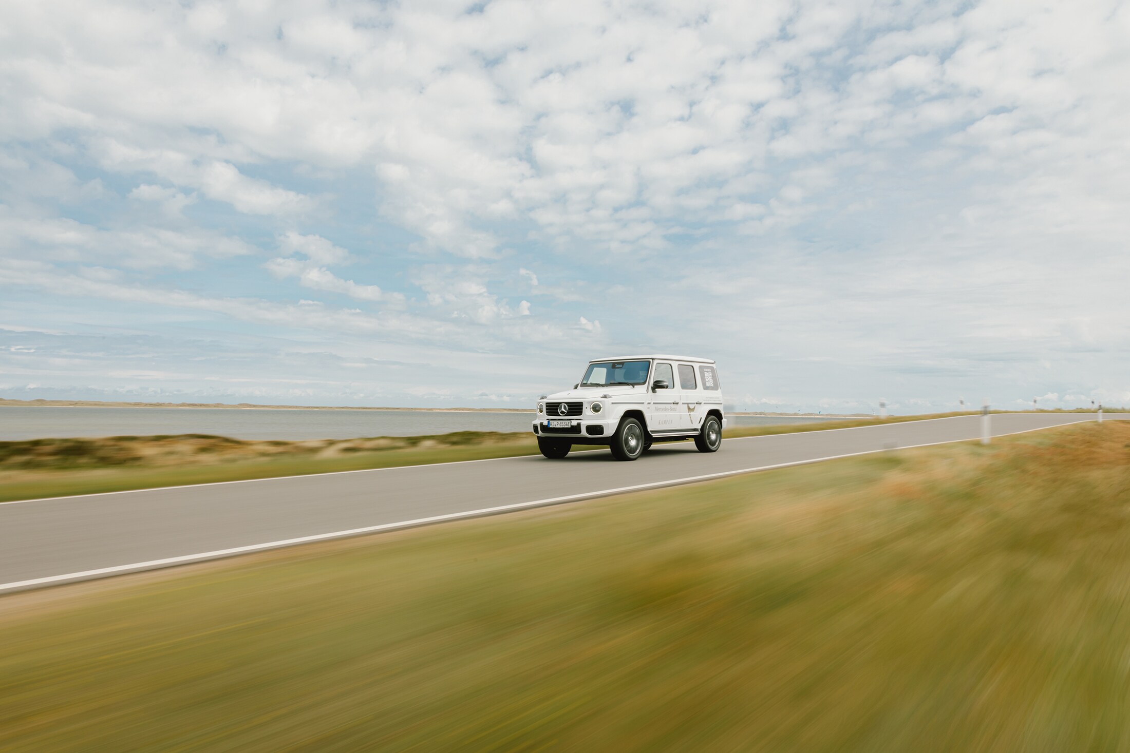Mercedes-Benz X Sylt | Mercedes-Benz Eine weiße Mercedes-Benz G-Klasse fährt über eine Landstraße am Wasser entlang.
