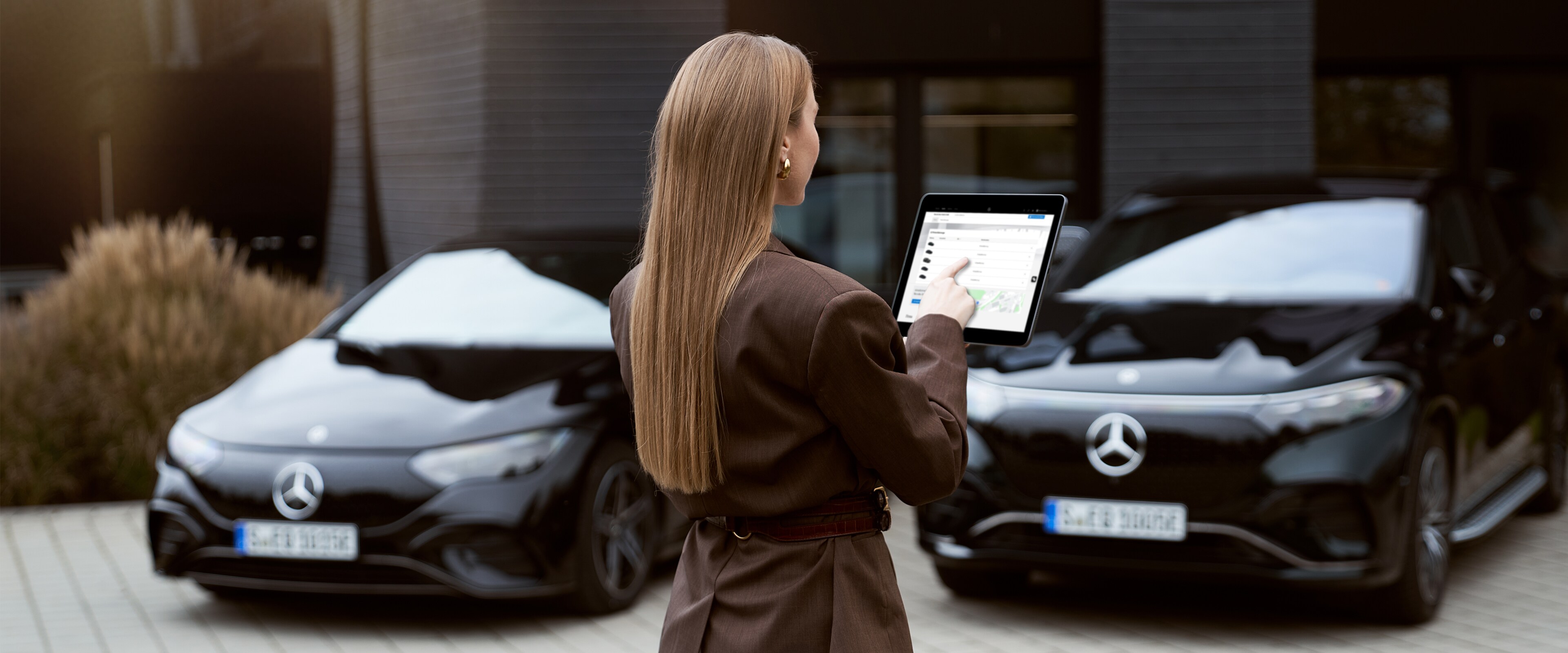 Flotten- und Geschäftskunden | Mercedes-Benz. Zwei schwarze Mercedes-Benz Fahrzeuge, parkend vor einem Gebäude. Eine Frau steht mit dem Rücken zum Bild und hat ein Tablett in der Hand-