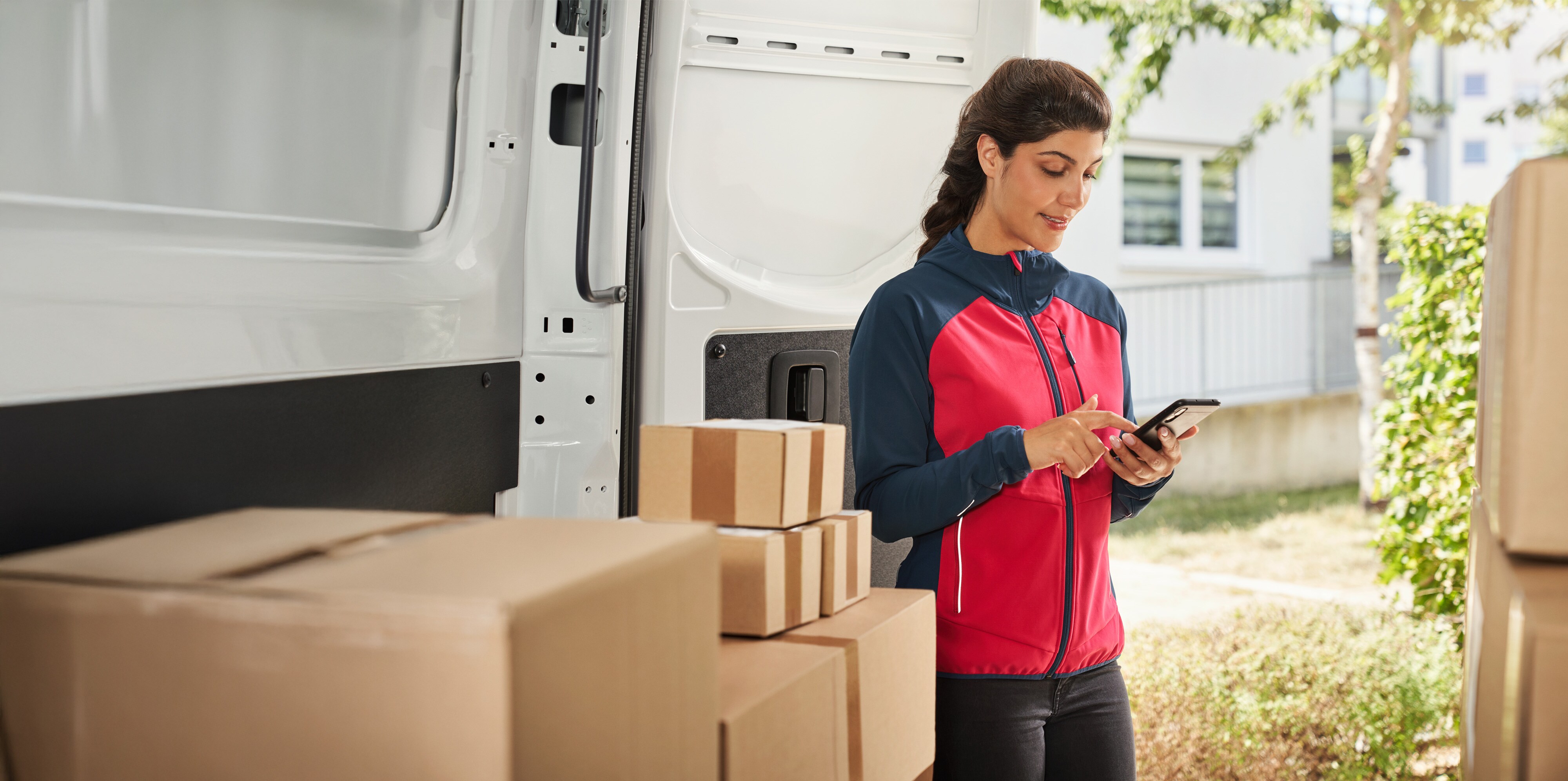 Customer Assistance Center | Mercedes-Benz Transporter. Zustellerin vor Lieferwagen mit Paketen nutzt Smartphone – professionelle Zustellung im Rahmen von Vans-Kundenservice.